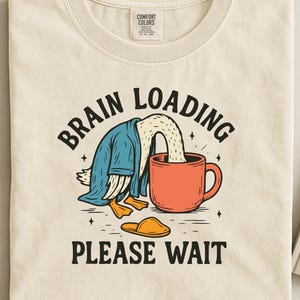 Puede incluir: Camiseta color crema con un gráfico humorístico. El diseño presenta un pato con la cabeza en una taza roja, vistiendo una bata azul. El texto dice "BRAIN LOADING PLEASE WAIT". Una zapatilla amarilla está en la parte inferior.