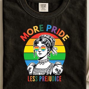 Op de afbeelding: Zwart T-shirt met een afbeelding van een vrouw met regenboog hartvormige bril, tegen een regenboog achtergrond. De tekst luidt "MORE PRIDE LESS PREJUDICE" in kleurrijke letters. Het shirt is gemaakt van een zacht materiaal.