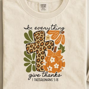 Op de afbeelding: Crèmekleurig T-shirt met een herfstthema. Het ontwerp bevat een kruis met een luipaardprint, een oranje pompoen en bloemelementen. De tekst luidt: "In everything give thanks 1 Thessalonians 5:18."