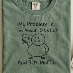 Op de afbeelding: Saliegroen t-shirt met de tekst "My Problem Is, I'm About 10% STUD And 90% Muffin." Een cartoonfiguur met een bierpul is ook afgebeeld. Het shirt heeft een ronde hals en een label "Comfort Colors."