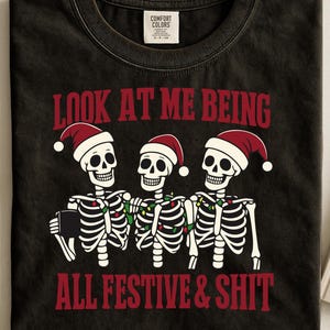 Op de afbeelding: Zwart t-shirt met een afbeelding van drie skeletten met kerstmutsen en kerstverlichting. De rode tekst luidt "LOOK AT ME BEING ALL FESTIVE & SHIT". Het shirt heeft een vintage uitstraling.