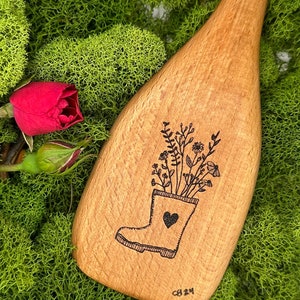 May include: A wooden spoon with a hand-drawn design of a boot filled with flowers. The boot has a heart on the side. The spoon is on a bed of green moss.
