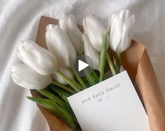 May include: A bouquet of white tulips wrapped in brown paper, with green stems and leaves. A white card with the text "and Baby Smith is ..." is tucked into the bouquet. The flowers rest on a white fabric surface.
