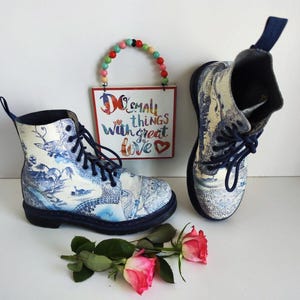 May include: A pair of blue and white patterned Dr. Martens boots with black laces. The boots feature a blue and white floral design with a blue and white bird on the side. The boots are sitting on a white surface with a sign that says "Do small things with great love" and a string of colourful beads.