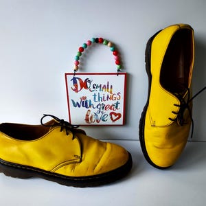 May include: A pair of yellow leather shoes with black laces. The shoes have a worn look and are sitting on a white surface. A sign with the text "Do small things with great love" is hanging above the shoes.