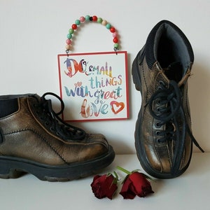Può includere: Due paia di stivali marroni in pelle con lacci neri. Gli stivali hanno un aspetto vissuto e sono appoggiati su una superficie bianca con un cartello rosso e bianco che dice "Do small things with great love".