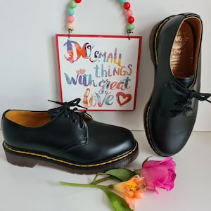 May include: A pair of black leather lace-up shoes with yellow stitching and a yellow sole. The shoes are on a white surface with a sign that says "Do small things with great love" and a pink flower.