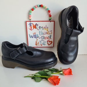Può includere: Un paio di scarpe Mary Jane in pelle nera con cinturino con fibbia e suola platform robusta. Le scarpe sono su una superficie bianca con un cartello che dice "Do small things with great love" e una collana di perline colorate.