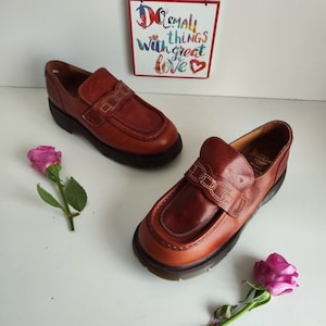 Può includere: Un paio di mocassini in pelle marrone con una suola spessa e un cinturino decorativo sul collo del piede. Le scarpe sono su una superficie bianca con rose rosa.