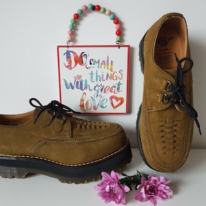 Può includere: Un paio di scarpe Dr. Martens in camoscio marrone con lacci neri e una spessa suola platform nera. Le scarpe hanno un design intrecciato sul davanti e un logo Dr. Martens all'interno della scarpa destra.