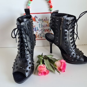 Può includere: Stivali neri in pelle con punta aperta, lacci e borchie. Gli stivali hanno una cerniera sul retro e un tacco alto.