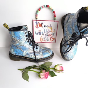 Pode incluir: Um par de botas azuis e brancas com padrão de pele de cobra e atacadores pretos. Uma placa com as palavras "Do small things with great love" está pendurada acima das botas. Duas rosas rosa e brancas e folhas verdes em primeiro plano.