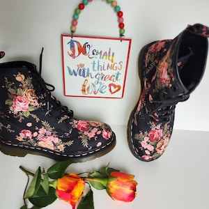 May include: Pair of black combat boots with a floral pattern of pink and yellow roses. The boots have black laces and a white stitched sole. A sign with the text "Do small things with great love" hangs above the boots.