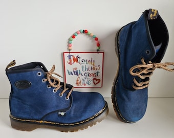 May include: A pair of blue canvas Dr. Martens boots with brown laces. The boots have a yellow logo on the side and a black rubber sole.
