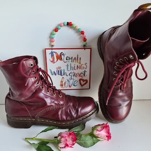 Può includere: Un paio di stivali stringati in pelle bordeaux con suola nera. Gli stivali sono usati e hanno un aspetto vissuto. Gli stivali sono appoggiati su una superficie bianca con un cartello che dice "Do small things with great love" davanti a loro.