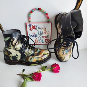 Peut inclure: Une paire de bottes en cuir noir avec un imprimé graphique coloré. Les bottes ont des lacets et une languette de traction jaune. Les bottes sont posées sur une surface blanche avec une petite pancarte qui dit "Faites de petites choses avec beaucoup d'amour."