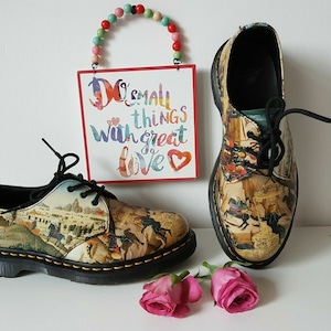 May include: Pair of leather shoes with a historical art print, black laces, and black soles. A sign with the text "Do small things with great love" hangs in the background. Two pink roses are in the foreground.