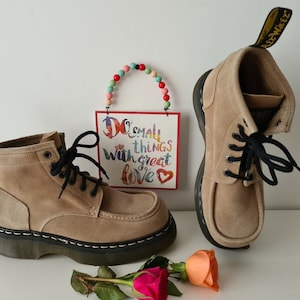 Può includere: Un paio di stivali da caviglia in camoscio color sabbia con lacci neri. Gli stivali hanno una suola nera spessa e un'etichetta gialla sul lato. Gli stivali sono appoggiati su una superficie bianca con un cartello che dice "Do small things with great love" e un cuore di perline.