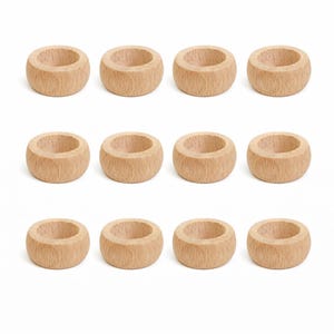 May include: Twelve wooden napkin rings arranged in a grid pattern. Each ring is a light brown color, with a rounded, cylindrical shape and a smooth, unfinished texture. The rings are evenly spaced against a white background.