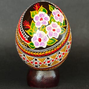 Op de afbeelding: Een decoratief ei met een zwarte basis, handbeschilderd met paarse bloemen, groene bladeren en complexe geometrische patronen in rood, geel en wit. Het ei rust op een donkerbruine ronde houten standaard en toont gedetailleerde volkskunstmotieven.
