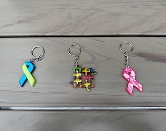 May include: Three keychains with different designs. The first keychain features a blue and yellow ribbon. The second keychain features a colorful puzzle piece. The third keychain features a pink ribbon.