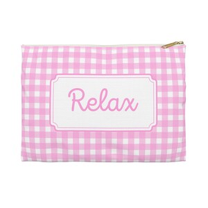 Puede incluir: Un estuche con cremallera con estampado de cuadros rosas y blancos con la palabra "Relax" en escritura rosa sobre una etiqueta rectangular blanca. El tirador de la cremallera es dorado.