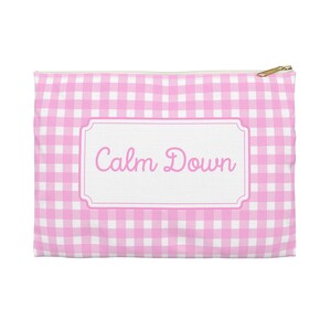 Puede incluir: Un estuche con cremallera con estampado de cuadros rosas y blancos con las palabras "Calm Down" en una fuente cursiva sobre una etiqueta rectangular blanca. La cremallera es dorada.