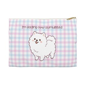 Puede incluir: Una bolsa con cremallera con un estampado de cuadros vichy en azul claro y rosa. La bolsa presenta una ilustración de dibujos animados de un perro blanco y esponjoso con el texto "I'm feeling overstimulated". La cremallera es dorada.