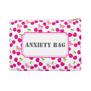 Könnte beinhalten: Weiße Reißverschlusstasche mit einem Muster aus rosa Kirschen und Schleifen. Die Worte "ANXIETY BAG" sind in Grau in einem weißen rechteckigen Rahmen aufgedruckt. Der Reißverschluss ist goldfarben.