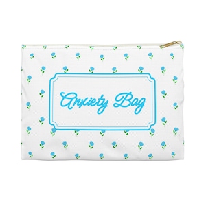 May include: White zippered pouch with a repeating pattern of small blue flowers and green leaves. The center features a light blue framed label with the words "Anxiety Bag" in a cursive font. The zipper is gold-colored.
