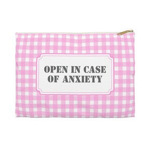 Könnte beinhalten: Ein rosa-weiß kariertes Reißverschlussetui mit dem Text "OPEN IN CASE OF ANXIETY" in einem weiß umrandeten Rechteck. Der Reißverschluss ist goldfarben.