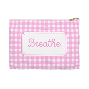 Puede incluir: Un estuche con cremallera con estampado de cuadros rosa y blanco con la palabra "Breathe" en escritura rosa. El texto está dentro de un marco rectangular blanco. La cremallera es dorada.