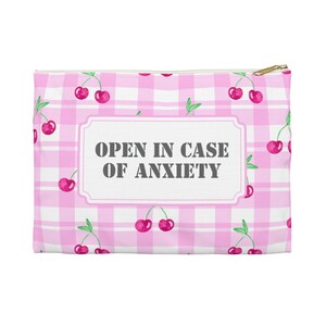 Könnte beinhalten: Ein rechteckiger, rosa-weiß karierter Reißverschlusspouch mit goldenem Reißverschluss. Der Beutel ist mit Kirschen und dem Text "OPEN IN CASE OF ANXIETY" in einem weißen Rahmen verziert.