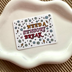 May include: White sticker with colorful text that reads "NEED A SENSORY BREAK" surrounded by small hearts, flowers, and lightning bolts.