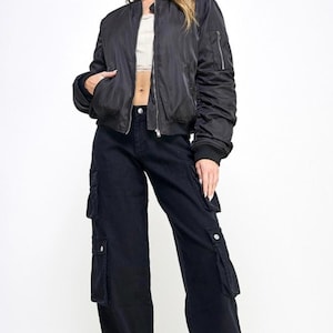 Könnte beinhalten: Eine schwarze Bomberjacke mit Reißverschluss und zwei Taschen. Die Jacke wird über einem weißen Crop-Top und einer schwarzen Cargohose getragen.