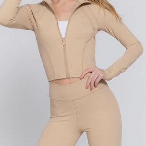 May include: A beige athletic outfit consisting of a zip-up jacket and leggings. The jacket has a high collar and long sleeves. The leggings are high-waisted. The outfit is made of a smooth, stretchy material, suitable for workouts or casual wear.