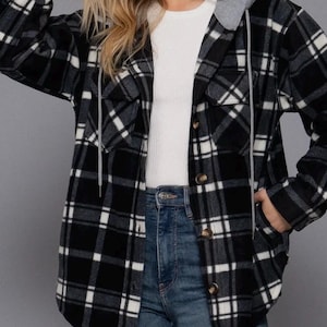 May include: A black and white checked flannel shirt jacket with a grey hood and drawstring. The jacket features two chest pockets and is buttoned. The model is wearing a white top and blue jeans.