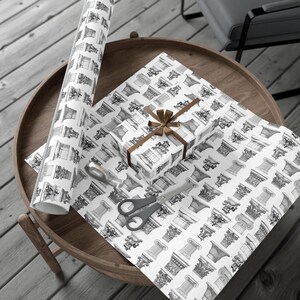 May include: A white gift wrap with a black and white pattern of architectural columns. A brown gift box with a brown ribbon is on top of the wrapping paper. A roll of the same wrapping paper is next to the gift box.