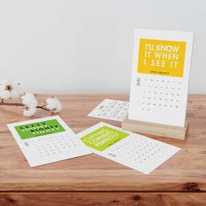May include: A desktop calendar with a wooden base, featuring the text "I'LL KNOW IT WHEN I SEE IT" in yellow. Additional calendar pages are scattered around, with text like "CUTTING CORNERS PERFECTLY" and "LEFTY LOOSEY RIGHTY".