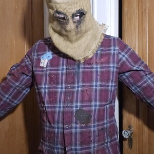 May include: A person wearing a brown burlap sack mask with eye holes and a red and blue plaid shirt. The shirt has several patches and appears to be distressed.