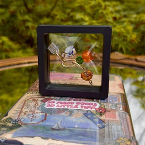 May include: A small, framed charm with a blue and white teacup, green and orange beads, and a gold chain. The charm is displayed on a book with the text "So many books, so little time".