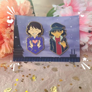 Op de afbeelding: Twee emaillepins met cartoonfiguren. De pin aan de linkerkant toont een personage met kort bruin haar, dat een paarse jas draagt en een roze hart vasthoudt. De pin aan de rechterkant toont een personage met bruin haar, dat een blauwe jas en een zwarte hoed draagt en een hart met een gat vasthoudt.