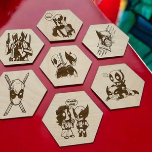 May include: Six hexagonal wooden coasters featuring different illustrations of Deadpool and Wolverine characters. The coasters are laser engraved with black and white images. The text on the coasters includes "Hello?", "I'm Marvel Jesus", and "Deadpool".