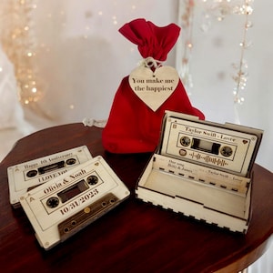 May include: Two wooden cassette tapes with personalized text, one with the text "Happy 4th Anniversary I LOVE YOU Olivia & Noah 10/31/2023" and the other with the text "Taylor Swift - Lover Our song Betty & James Taylor Swift - Lover". A red velvet bag with a heart-shaped tag that reads "You make me the happiest" is also in the image.
