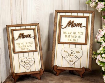 Op de afbeelding: Twee houten frames met een witte achtergrond en gouden letters. Het bovenste frame zegt "Mam Je Bent Het Stukje Dat Ons Samenhoudt". Het onderste frame heeft een hartvormige puzzel met de namen "Peter, Kate, Mam, Pap, Nick, Gabrial en Liam" op elk stuk.