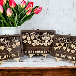 May include: Three wooden plaques with a tree branch design and hearts hanging from the branches. Each heart has a name written on it. The plaques read "Miller Family", "Merry Christmas", and "We Are Family".