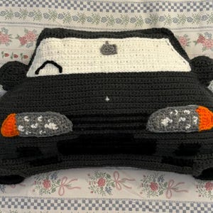 Puede incluir: Un cojín hecho a mano con forma de coche de ganchillo en gris oscuro, blanco y naranja. El cojín presenta un parabrisas, faros y espejos laterales. El diseño del coche es caprichoso y adecuado para la habitación de un niño o como objeto decorativo.