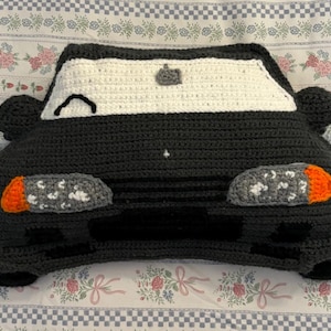 Peut inclure: Un coussin fait à la main en forme de voiture au crochet, de couleur gris foncé, blanc et orange. Le coussin présente un pare-brise, des phares et des rétroviseurs. Le design de la voiture est fantaisiste et convient à une chambre d'enfant ou comme objet de décoration.