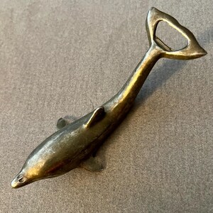 Könnte beinhalten: Ein bronzefarbener Flaschenöffner in Form eines Delfins. Der Flaschenöffner besteht aus Metall und hat ein Vintage-Aussehen. Der Schwanz des Delfins bildet den Flaschenöffner, und der Körper hat Flossen. Es ist ein dekorativer Artikel.