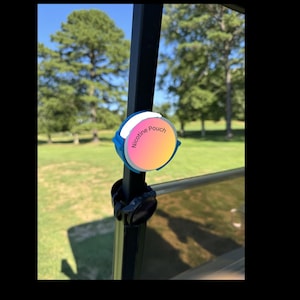 May include: A blue and white Nicotine Pouch container with a gradient pink and yellow center, mounted on a black bar. The text "Nicotine Pouch" is printed on the container. The background shows a green field and trees.